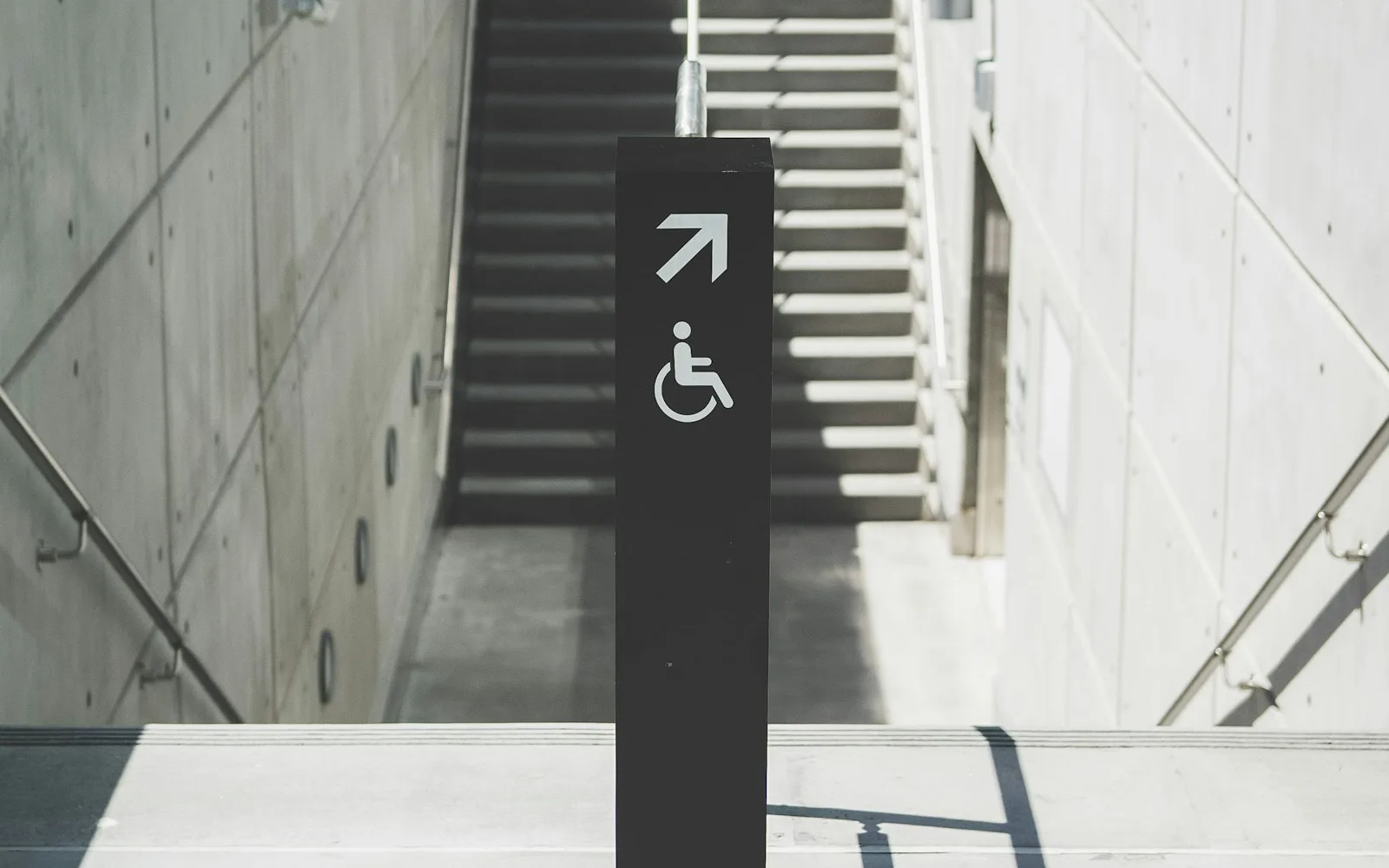 Cartello di accessibilità con freccia davanti a una rampa di scale in cemento.