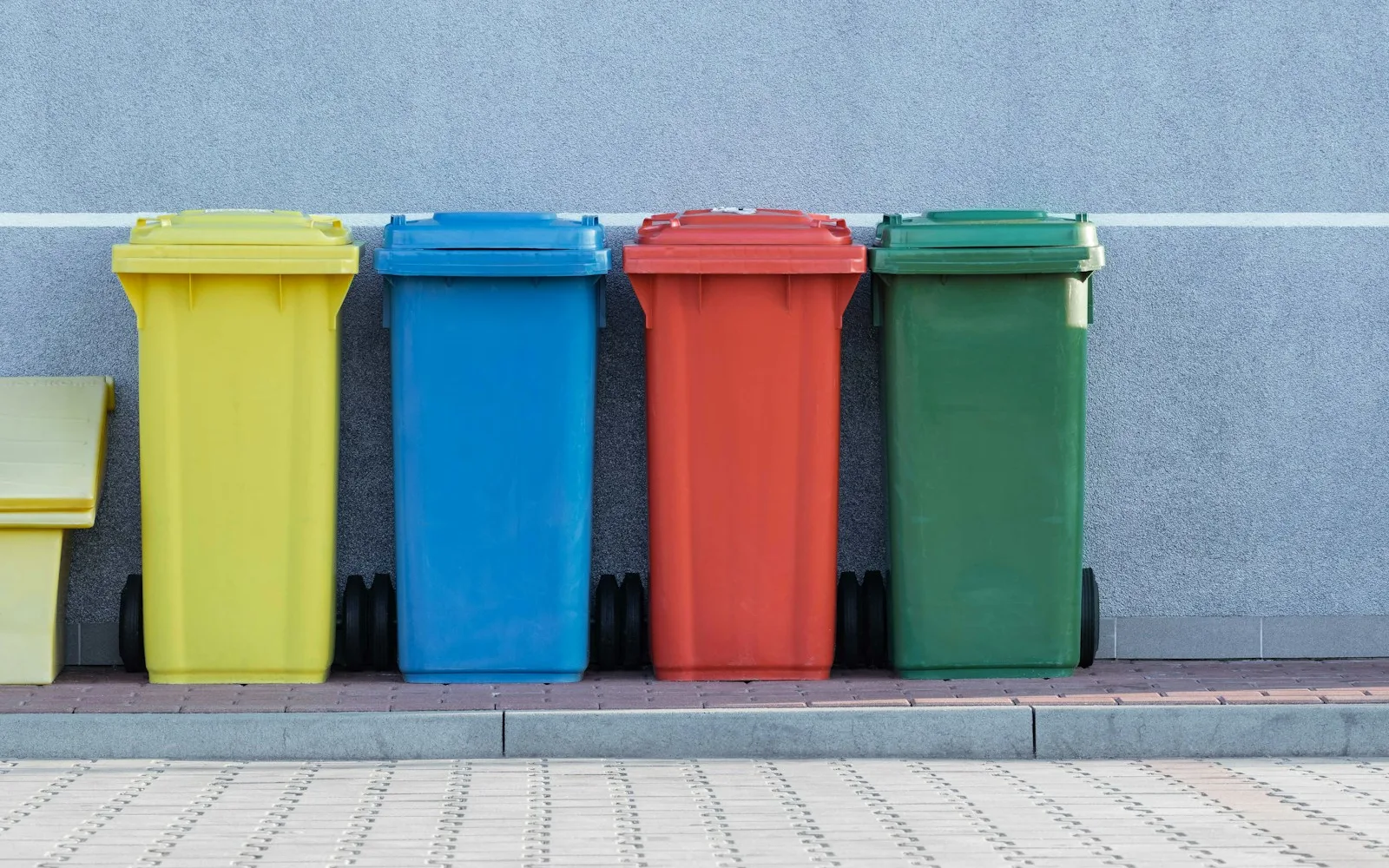 Fila di bidoni colorati per la raccolta differenziata addossati al muro di un edificio.