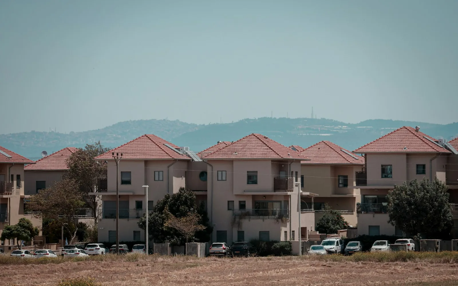 Fila di edifici residenziali bassi con tetti in coppi, balconi e parcheggio comune davanti.