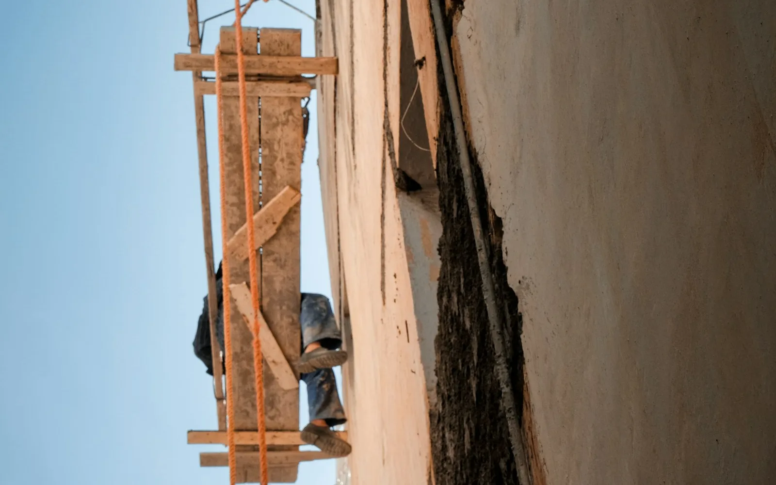Operaio su un ponteggio artigianale addossato alla facciata intonacata di un edificio.