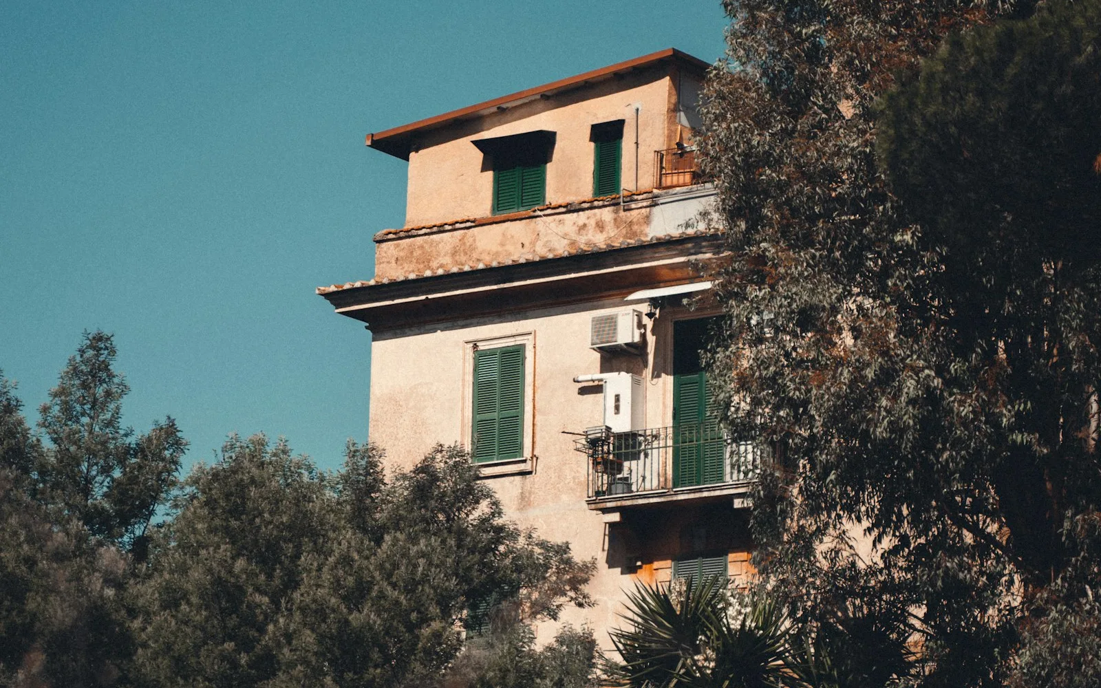 Palazzina residenziale italiana con persiane verdi e alberi ad alto fusto cresciuti a ridosso della facciata.