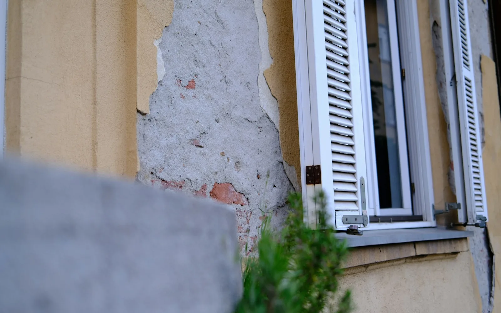 Intonaco staccato fino al mattone sulla facciata di un edificio residenziale, accanto a una finestra con persiane bianche.