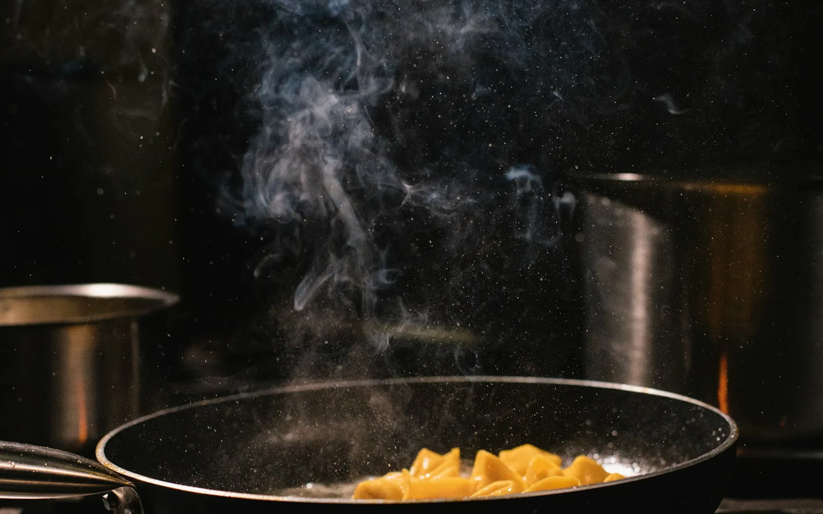 Padella su un fornello con vapori di cottura che salgono, in penombra.
