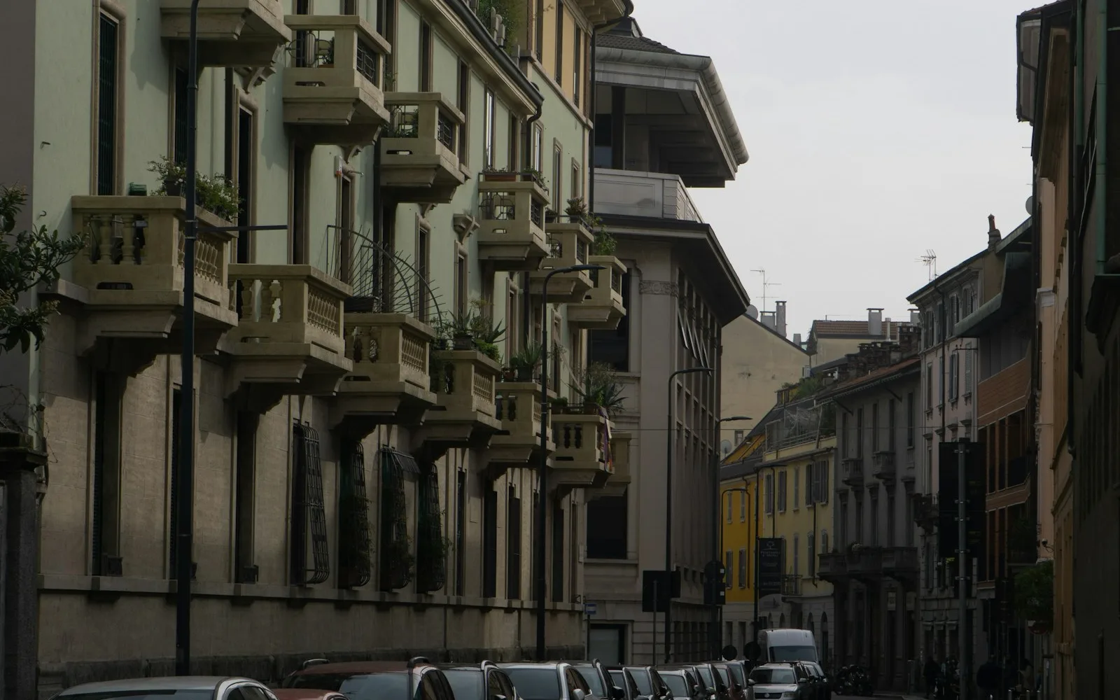 Via milanese con palazzine dei primi del Novecento, balconi fioriti e auto in sosta.