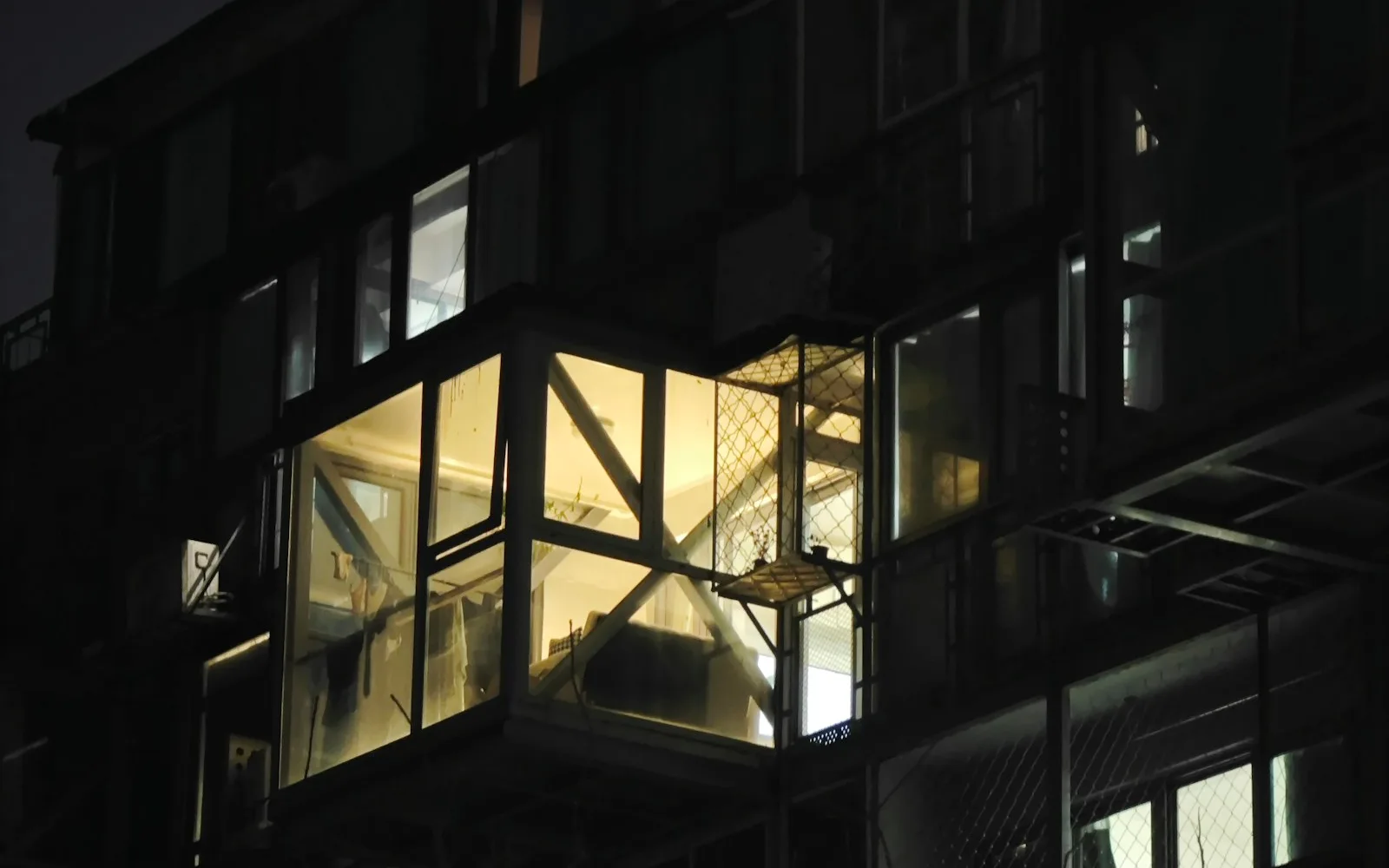 Balcone chiuso con vetrate illuminato dall'interno sulla facciata scura di un edificio residenziale di sera.
